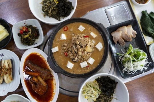 위에서 본 한 상 — 가운데 청국장 뚝배기, 둘레에 고기 접시·붉은 양념 생선조림·나물 반찬